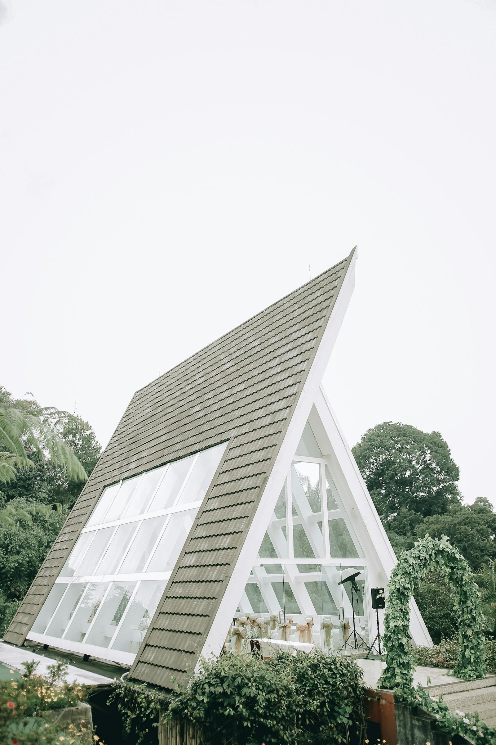 Exterior of contemporary geometric building with windows near green archway and plants near forest under cloudless sky in daytime in summer